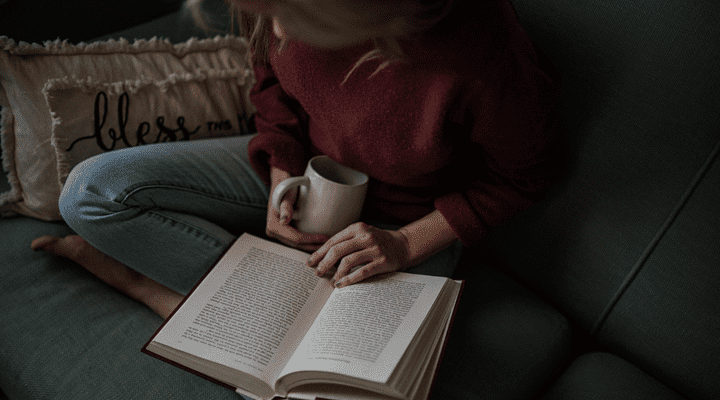 A woman sitting on a sofa reading a book with a cup of coffee