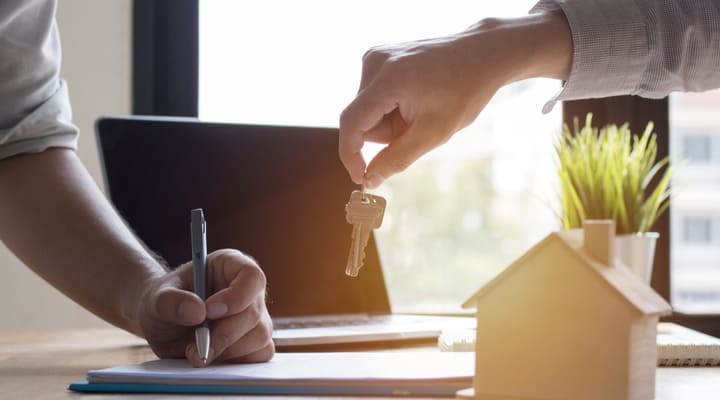 A man signing paperwork as someone holds out a house key.