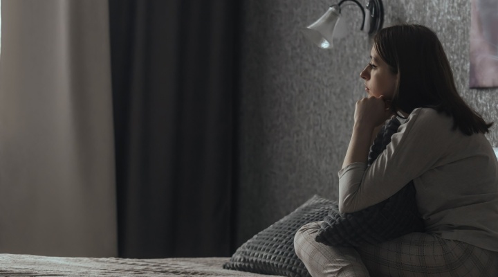 A woman sits in a dark bedroom looking sad.